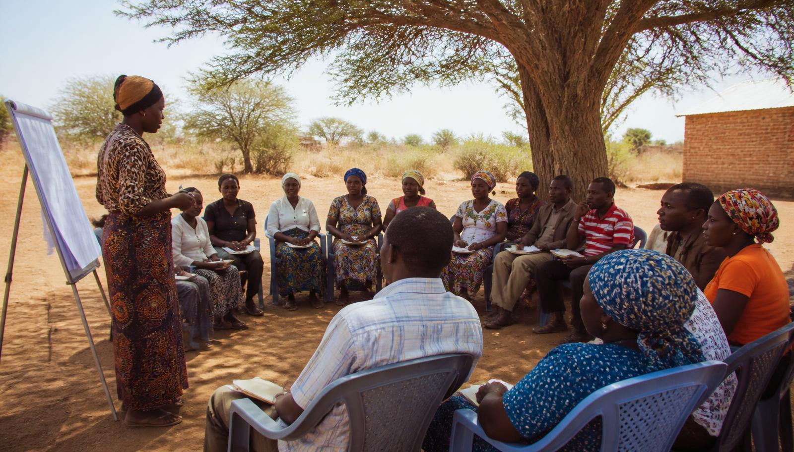 Parents and caregivers seated in a semicircle under an acacia tree while a facilitator presents at a flip chart.
