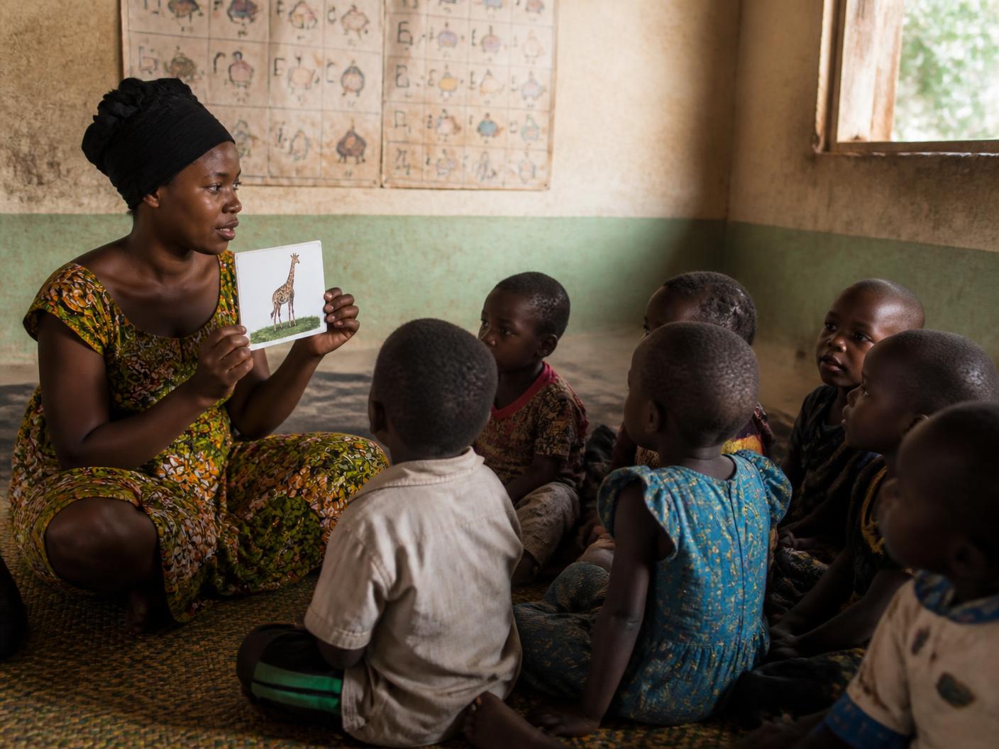 A community early-learning facilitator holds up a picture card for a group of young children seated on a mat.