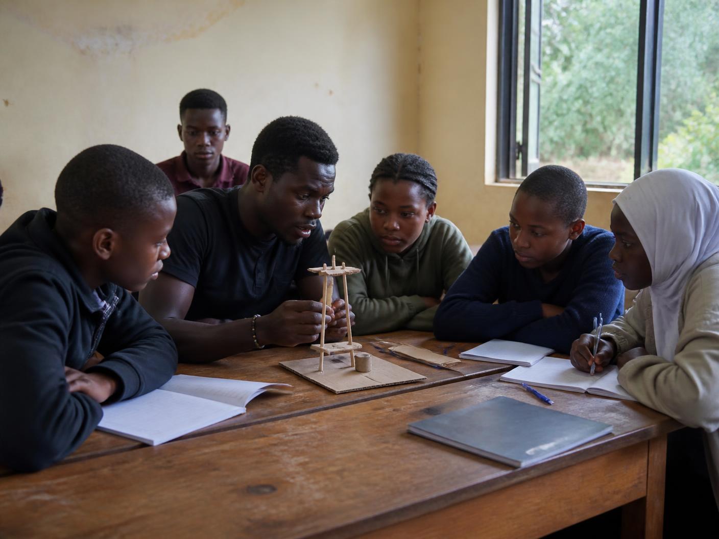 A young mentor and five teenagers examining a handmade prototype around a wooden table with notebooks open.
